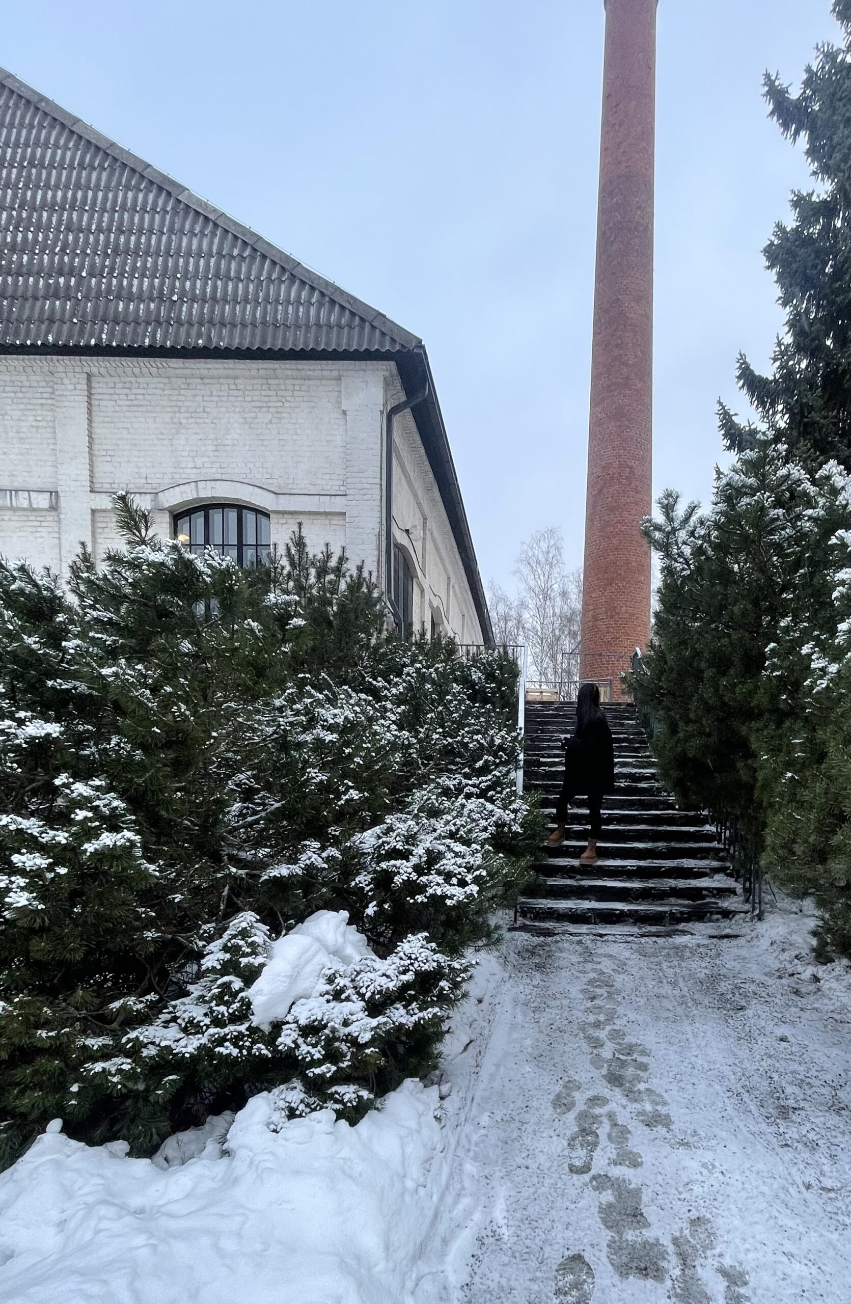 Henkilö seisoo lumisilla rappusilla Riihimäen lasimuseon edustalla. Taustalla näkyy korkea tiilestä tehty tehtaanpiippu.